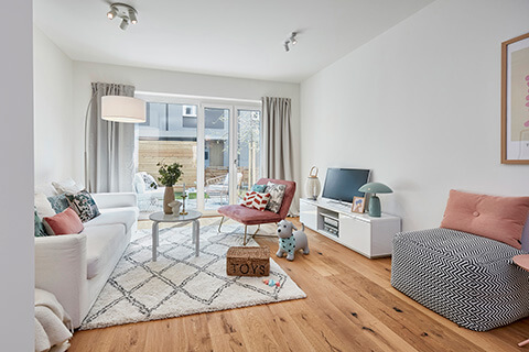 Wohnzimmer mit Sofa links und Fernseher rechts sowie Blick aus dem Fenster der zeiten Musterwohnung des Neubauprojekts Constanze im ConstantinQuartier.
